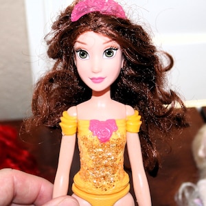 May include: A doll with brown hair and green eyes, wearing a yellow and gold glittery dress with a pink rose detail. The doll has a pink tiara and is held by a hand.