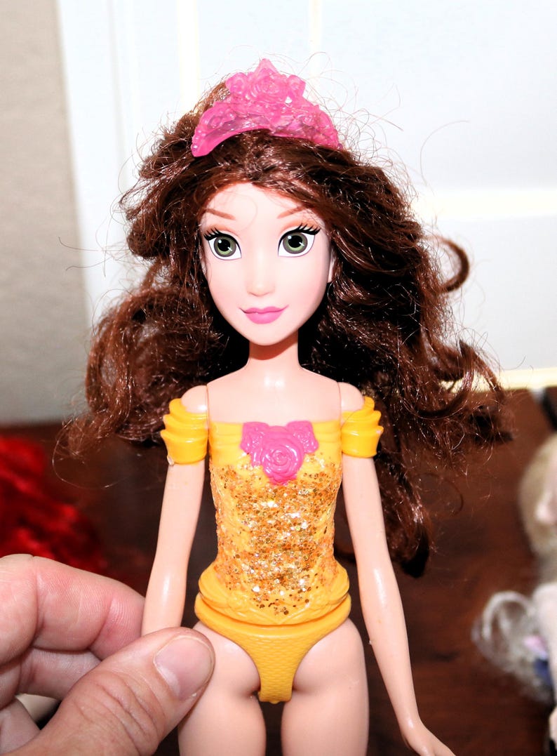 May include: A doll with brown hair and green eyes, wearing a yellow and gold glittery dress with a pink rose detail. The doll has a pink tiara and is held by a hand.