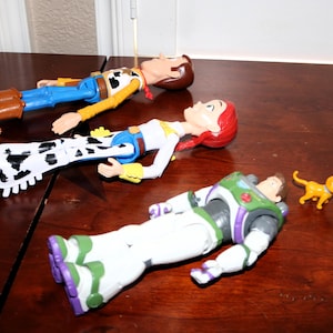 Puede incluir: Tres figuras de acci&oacute;n de los personajes de Toy Story: Woody, Jessie y Buzz Lightyear, junto con una peque&ntilde;a figura de gato naranja, est&aacute;n dispuestas sobre una superficie de madera oscura. Woody viste un atuendo de vaquero, Jessie un atuendo de vaquera y Buzz un traje espacial.
