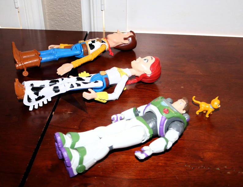 Puede incluir: Tres figuras de acci&oacute;n de los personajes de Toy Story: Woody, Jessie y Buzz Lightyear, junto con una peque&ntilde;a figura de gato naranja, est&aacute;n dispuestas sobre una superficie de madera oscura. Woody viste un atuendo de vaquero, Jessie un atuendo de vaquera y Buzz un traje espacial.
