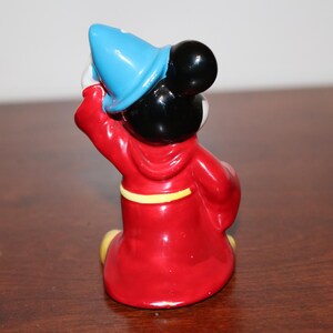 May include: A ceramic figurine of Mickey Mouse dressed as a sorcerer. He is wearing a red robe with a yellow sash, a blue pointed hat, and black shoes. He is holding a wand in his right hand.