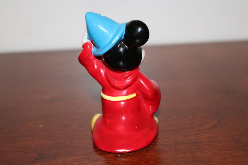 May include: A ceramic figurine of Mickey Mouse dressed as a sorcerer. He is wearing a red robe with a yellow sash, a blue pointed hat, and black shoes. He is holding a wand in his right hand.