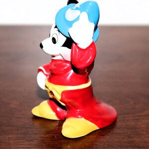 May include: A ceramic figurine of Mickey Mouse dressed as a sorcerer. He is wearing a red robe, a blue hat, and yellow shoes. He is holding his hat in his right hand and has a mischievous expression on his face.