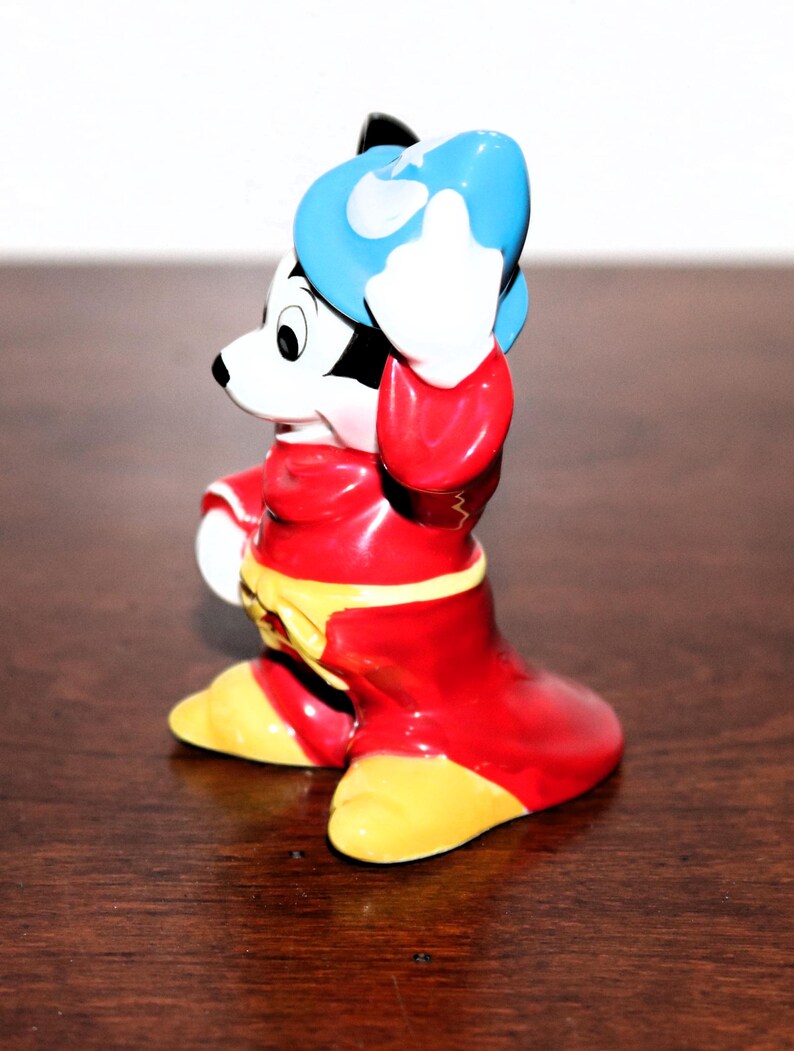 May include: A ceramic figurine of Mickey Mouse dressed as a sorcerer. He is wearing a red robe, a blue hat, and yellow shoes. He is holding his hat in his right hand and has a mischievous expression on his face.