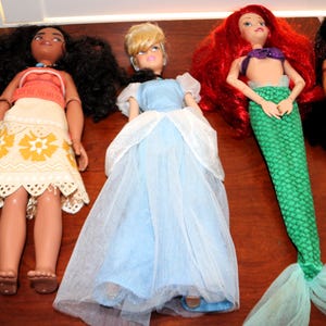 May include: Four fashion dolls arranged in a row. The first has long dark curly hair, a coral top, and a cream skirt with yellow floral patterns. The second has blonde hair and a light blue dress with a white tulle overlay. The third has long red hair and a green mermaid tail. The fourth has long black hair and white underwear.