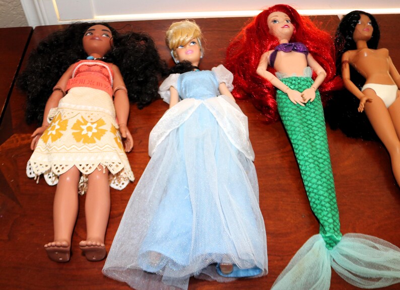 May include: Four fashion dolls arranged in a row. The first has long dark curly hair, a coral top, and a cream skirt with yellow floral patterns. The second has blonde hair and a light blue dress with a white tulle overlay. The third has long red hair and a green mermaid tail. The fourth has long black hair and white underwear.