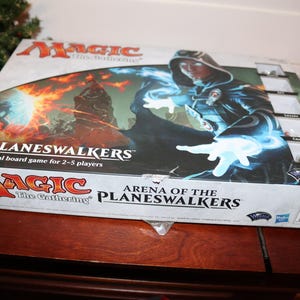 May include: A Magic: The Gathering board game box with a blue and white image of a planeswalker. The box is titled "Arena of the Planeswalkers" and features the Magic: The Gathering logo.
