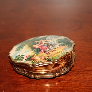 May include: A gold-toned compact with a white and green floral design featuring a couple dancing. The compact has a scalloped edge and a hinged lid.