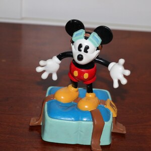 May include: A Mickey Mouse figurine stands on a blue box with brown straps. Mickey is wearing red shorts, yellow shoes, and white gloves. His face is white with black eyes and ears. He has a blue and white hat.