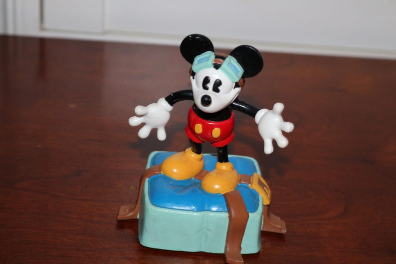 May include: A Mickey Mouse figurine stands on a blue box with brown straps. Mickey is wearing red shorts, yellow shoes, and white gloves. His face is white with black eyes and ears. He has a blue and white hat.