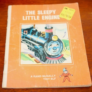 May include: Vintage children's book titled "The Sleepy Little Engine" with an illustration of a blue train on the cover. The book has an orange cover with the text "A Rand McNally Tiny Elf". A price tag is visible in the upper right corner.