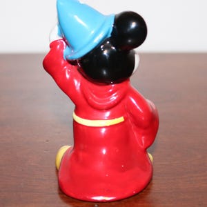 May include: A ceramic figurine of Mickey Mouse dressed as a sorcerer. He is wearing a red robe with a yellow sash, a blue pointed hat, and black shoes. He is holding his hat in his right hand and his left hand is at his side.
