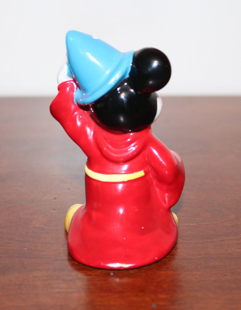 May include: A ceramic figurine of Mickey Mouse dressed as a sorcerer. He is wearing a red robe with a yellow sash, a blue pointed hat, and black shoes. He is holding his hat in his right hand and his left hand is at his side.