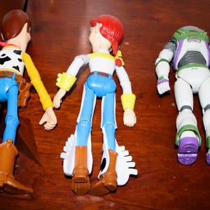 Puede incluir: Tres figuras de acci&oacute;n de personajes de Toy Story: Woody, Jessie y Buzz Lightyear, junto con una peque&ntilde;a figura de gato amarillo. Woody lleva sombrero de vaquero, chaleco y botas. Jessie tiene pelo rojo y un atuendo de vaquera. Buzz lleva un traje espacial.