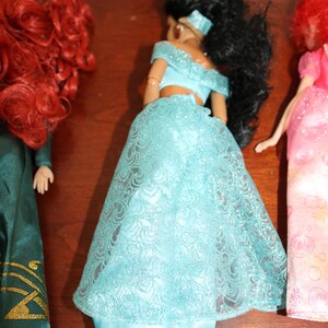 Puede incluir: Se muestran tres mu&ntilde;ecas, cada una con caracter&iacute;sticas distintas. La mu&ntilde;eca central viste un top azul claro con hombros descubiertos y una falda a juego con una superposici&oacute;n de encaje. La mu&ntilde;eca de la izquierda tiene cabello rizado rojo y un vestido verde. La mu&ntilde;eca de la derecha tiene cabello rojo y un vestido rosa.