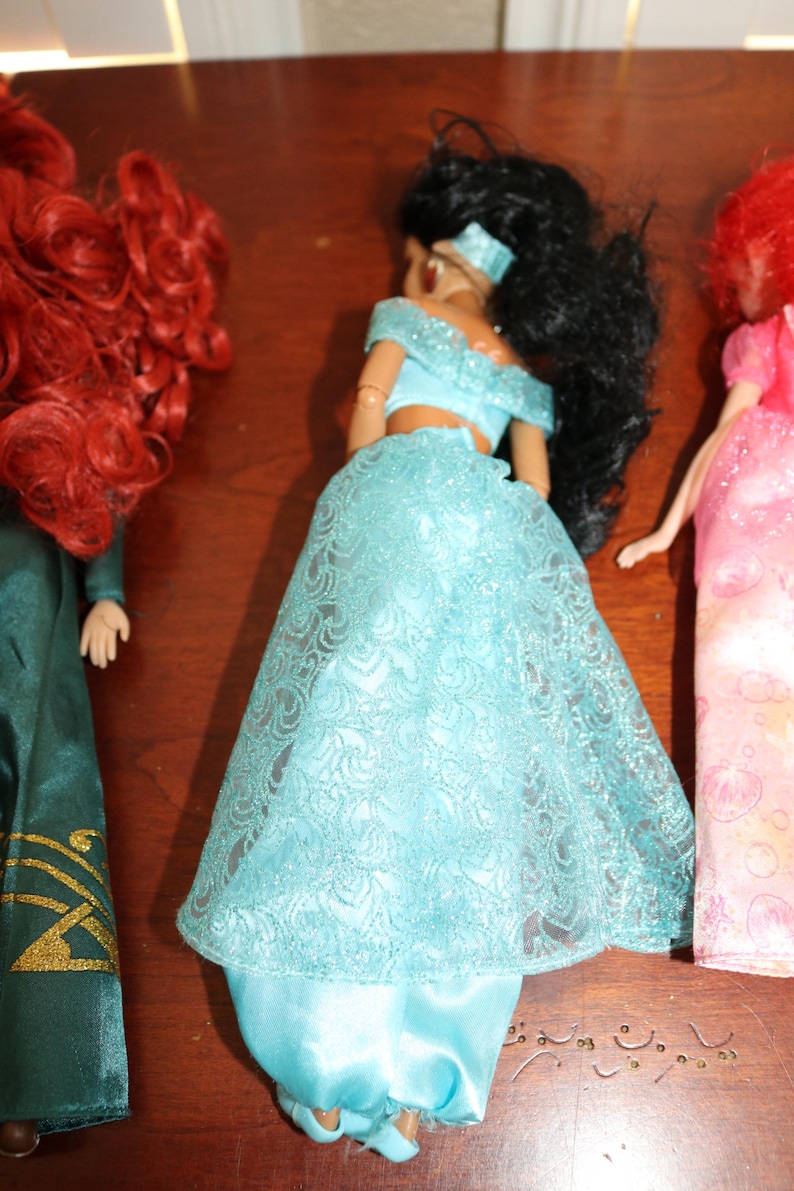 Puede incluir: Se muestran tres mu&ntilde;ecas, cada una con caracter&iacute;sticas distintas. La mu&ntilde;eca central viste un top azul claro con hombros descubiertos y una falda a juego con una superposici&oacute;n de encaje. La mu&ntilde;eca de la izquierda tiene cabello rizado rojo y un vestido verde. La mu&ntilde;eca de la derecha tiene cabello rojo y un vestido rosa.
