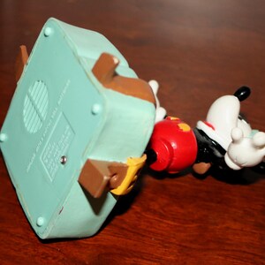 May include: A Mickey Mouse figurine, featuring a red shorts, black jacket, white gloves, and a white face with black ears. The figurine is lying on its side, with a light blue base and brown accents.