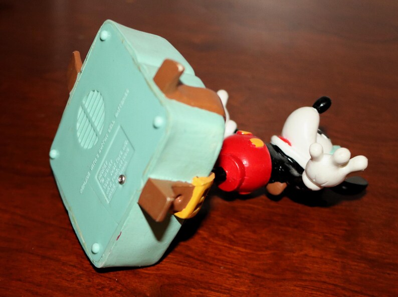 May include: A Mickey Mouse figurine, featuring a red shorts, black jacket, white gloves, and a white face with black ears. The figurine is lying on its side, with a light blue base and brown accents.