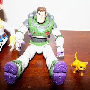 Puede incluir: Figura de acci&oacute;n de Buzz Lightyear con un traje espacial verde y blanco, detalles morados y una figura de gato amarillo. La figura de Buzz Lightyear tiene una cabeza marr&oacute;n y un casco verde. La figura del gato es naranja con ojos negros.