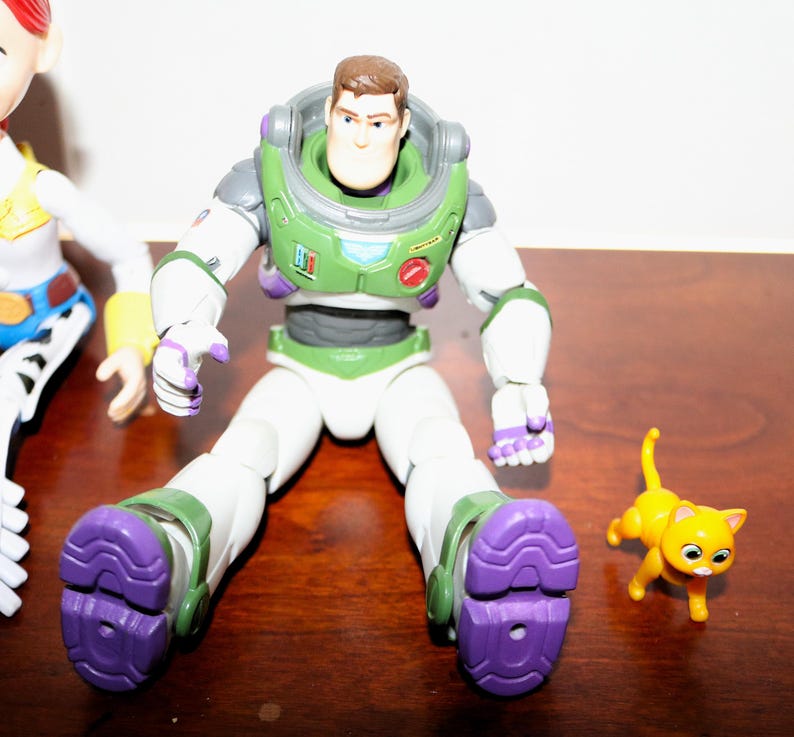 Puede incluir: Figura de acci&oacute;n de Buzz Lightyear con un traje espacial verde y blanco, detalles morados y una figura de gato amarillo. La figura de Buzz Lightyear tiene una cabeza marr&oacute;n y un casco verde. La figura del gato es naranja con ojos negros.