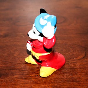 May include: A ceramic figurine of Mickey Mouse dressed as a sorcerer. He is wearing a red robe, a blue hat with a white star, and yellow shoes. He is holding his hat in his right hand and has a mischievous expression on his face.