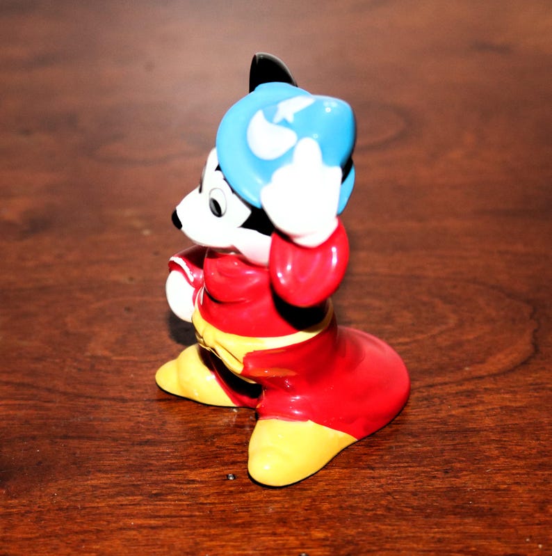 May include: A ceramic figurine of Mickey Mouse dressed as a sorcerer. He is wearing a red robe, a blue hat with a white star, and yellow shoes. He is holding his hat in his right hand and has a mischievous expression on his face.
