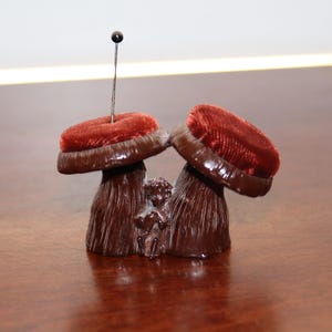May include: A vintage pin cushion with a dark brown base and two mushroom-shaped cushions topped with red velvet. A small cherub figure is between the mushrooms. A pin with a black head is inserted into one cushion.