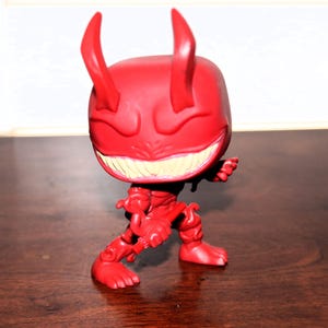 May include: A red vinyl figure of a devil-like character with large horns, a wide grin revealing yellow teeth, and a muscular physique. The figure is posed in a dynamic stance, with its arms and legs slightly bent.