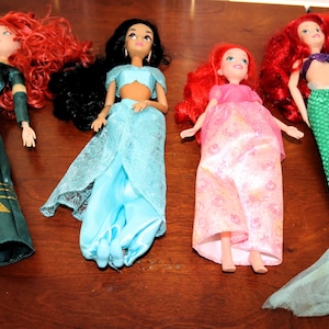 Puede incluir: Se muestran cuatro mu&ntilde;ecas, cada una representando un personaje diferente. Las mu&ntilde;ecas tienen el pelo largo de varios colores, incluyendo rojo y negro. Visten trajes coloridos, incluyendo un vestido verde, pantalones azules y una cola de sirena.
