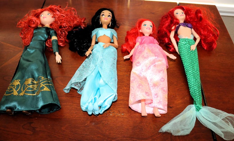 Puede incluir: Se muestran cuatro mu&ntilde;ecas, cada una representando un personaje diferente. Las mu&ntilde;ecas tienen el pelo largo de varios colores, incluyendo rojo y negro. Visten trajes coloridos, incluyendo un vestido verde, pantalones azules y una cola de sirena.