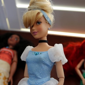 May include: A blonde doll with an updo hairstyle wearing a light blue dress with white sheer puff sleeves, a black choker, and small blue stud earrings. The bodice features a central blue gem accent and a subtle glittery finish on the fabric.