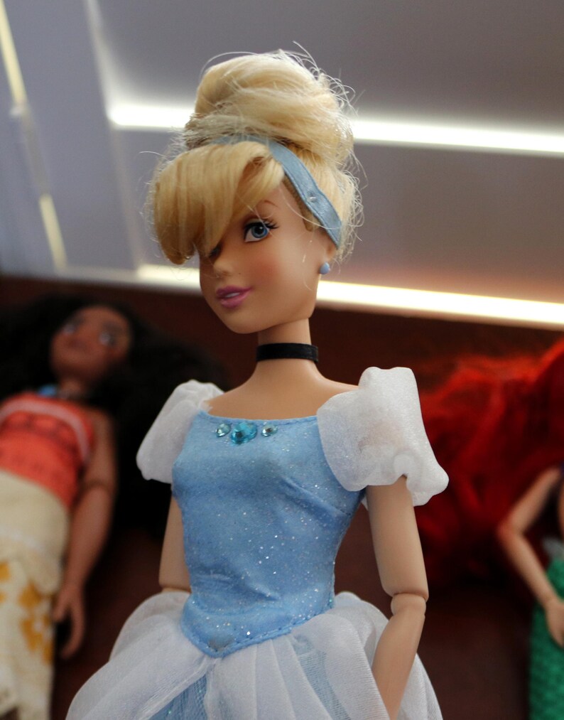 May include: A blonde doll with an updo hairstyle wearing a light blue dress with white sheer puff sleeves, a black choker, and small blue stud earrings. The bodice features a central blue gem accent and a subtle glittery finish on the fabric.
