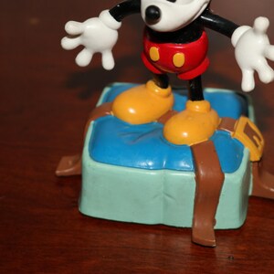 May include: A Mickey Mouse figurine stands on a light blue and green box with brown straps. Mickey is wearing red shorts, yellow shoes, and white gloves. The figurine is a collectible item.