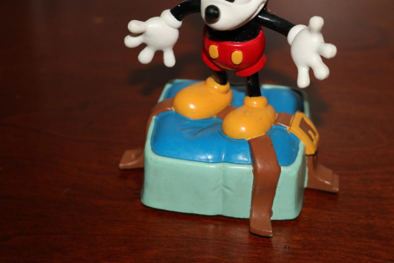 May include: A Mickey Mouse figurine stands on a light blue and green box with brown straps. Mickey is wearing red shorts, yellow shoes, and white gloves. The figurine is a collectible item.