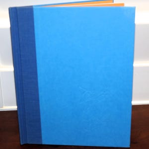 May include: A hardcover book with a bright blue cover and a dark blue spine. The pages have orange edges. The book is standing upright on a wooden surface.