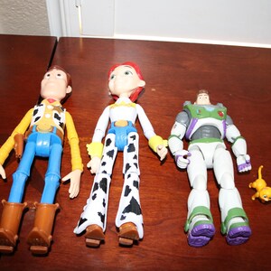 Puede incluir: Tres figuras de acci&oacute;n de Toy Story: Woody con un atuendo de vaquero, Jessie con un atuendo de vaquera y Buzz Lightyear con un traje espacial. Tambi&eacute;n hay una peque&ntilde;a figura de gato amarillo. Los juguetes est&aacute;n sobre una superficie marr&oacute;n.