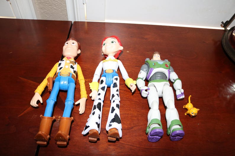 Puede incluir: Tres figuras de acci&oacute;n de Toy Story: Woody con un atuendo de vaquero, Jessie con un atuendo de vaquera y Buzz Lightyear con un traje espacial. Tambi&eacute;n hay una peque&ntilde;a figura de gato amarillo. Los juguetes est&aacute;n sobre una superficie marr&oacute;n.
