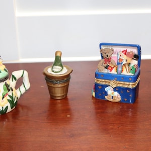 May include: A collection of decorative trinket boxes. Includes a teapot, a champagne bottle in a bucket, a blue box with a teddy bear, and a bunny in a basket. The boxes are painted with various colors and designs.