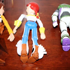 Puede incluir: Tres figuras de acci&oacute;n de los personajes de Toy Story: Woody, Jessie y Buzz Lightyear, junto con una peque&ntilde;a figura de gato naranja. Woody lleva sombrero de vaquero, chaleco y botas. Jessie tiene un sombrero rojo y pantalones azules. Buzz lleva un traje espacial blanco y morado.