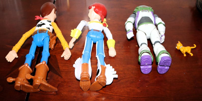 Puede incluir: Tres figuras de acci&oacute;n de los personajes de Toy Story: Woody, Jessie y Buzz Lightyear, junto con una peque&ntilde;a figura de gato naranja. Woody lleva sombrero de vaquero, chaleco y botas. Jessie tiene un sombrero rojo y pantalones azules. Buzz lleva un traje espacial blanco y morado.