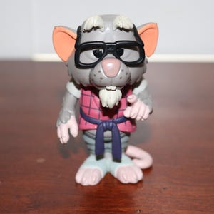 May include: A gray anthropomorphic rat figurine wearing black-rimmed glasses, a red and black plaid shirt, and a dark purple belt. The character has light-colored tufts of hair on its head and chin, large pink-lined ears, and a pink tail.