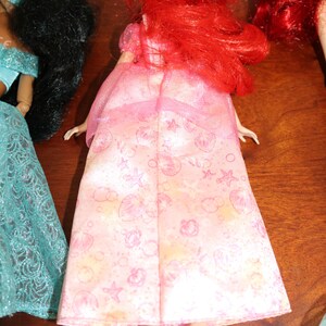 Puede incluir: Un vestido de mu&ntilde;eca rosa con un estampado de conchas, estrellas y nubes. La mu&ntilde;eca tiene el pelo rojo y lleva un vestido rosa. La mu&ntilde;eca forma parte de una colecci&oacute;n de mu&ntilde;ecas.
