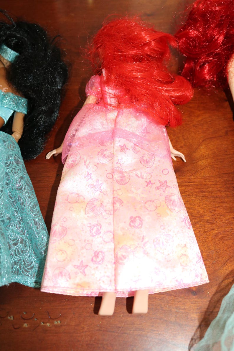 Puede incluir: Un vestido de mu&ntilde;eca rosa con un estampado de conchas, estrellas y nubes. La mu&ntilde;eca tiene el pelo rojo y lleva un vestido rosa. La mu&ntilde;eca forma parte de una colecci&oacute;n de mu&ntilde;ecas.