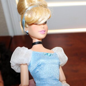 May include: A doll with blonde hair styled in an updo with a light blue headband. The doll wears a light blue bodice with a shimmering finish, white sheer puff sleeves, a black choker, and small blue stud earrings. The outfit features a blue gem.