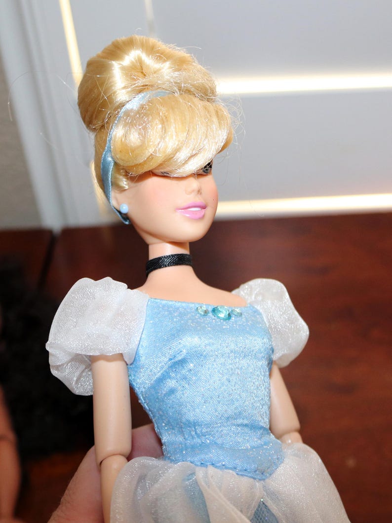 May include: A doll with blonde hair styled in an updo with a light blue headband. The doll wears a light blue bodice with a shimmering finish, white sheer puff sleeves, a black choker, and small blue stud earrings. The outfit features a blue gem.