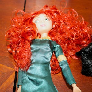 Puede incluir: Mu&ntilde;eca con cabello rizado rojo brillante y ojos verdes. La mu&ntilde;eca viste un vestido verde azulado con mangas largas y adornos dorados. La mu&ntilde;eca tiene la piel clara y una ligera sonrisa. Est&aacute; sobre una superficie de madera.
