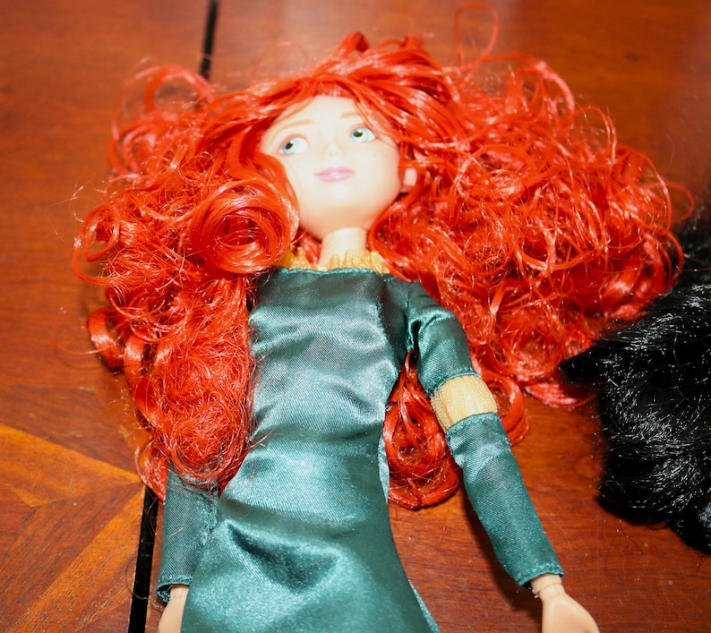Puede incluir: Mu&ntilde;eca con cabello rizado rojo brillante y ojos verdes. La mu&ntilde;eca viste un vestido verde azulado con mangas largas y adornos dorados. La mu&ntilde;eca tiene la piel clara y una ligera sonrisa. Est&aacute; sobre una superficie de madera.