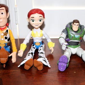 Puede incluir: Tres figuras de personajes de Toy Story: Woody, Jessie y Buzz Lightyear, junto con una peque&ntilde;a figura de gato naranja. Woody viste camisa amarilla, vaqueros azules y sombrero de vaquero. Jessie tiene un atuendo de vaquera. Buzz lleva un traje espacial.