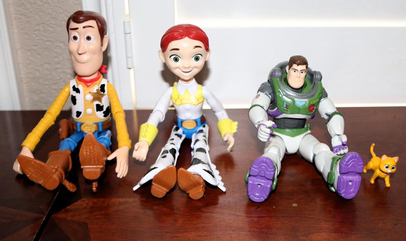 Puede incluir: Tres figuras de personajes de Toy Story: Woody, Jessie y Buzz Lightyear, junto con una peque&ntilde;a figura de gato naranja. Woody viste camisa amarilla, vaqueros azules y sombrero de vaquero. Jessie tiene un atuendo de vaquera. Buzz lleva un traje espacial.