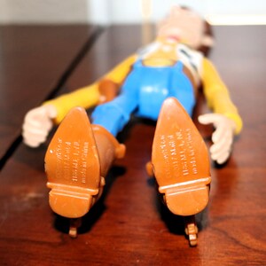 Puede incluir: Figura de juguete de un vaquero tumbado boca arriba. La figura lleva una camisa amarilla, un peto azul y botas marrones. Se ven las suelas de las botas, con el texto "&copy;2017 Mattel 1186 MJ, 1, NC, made in China."